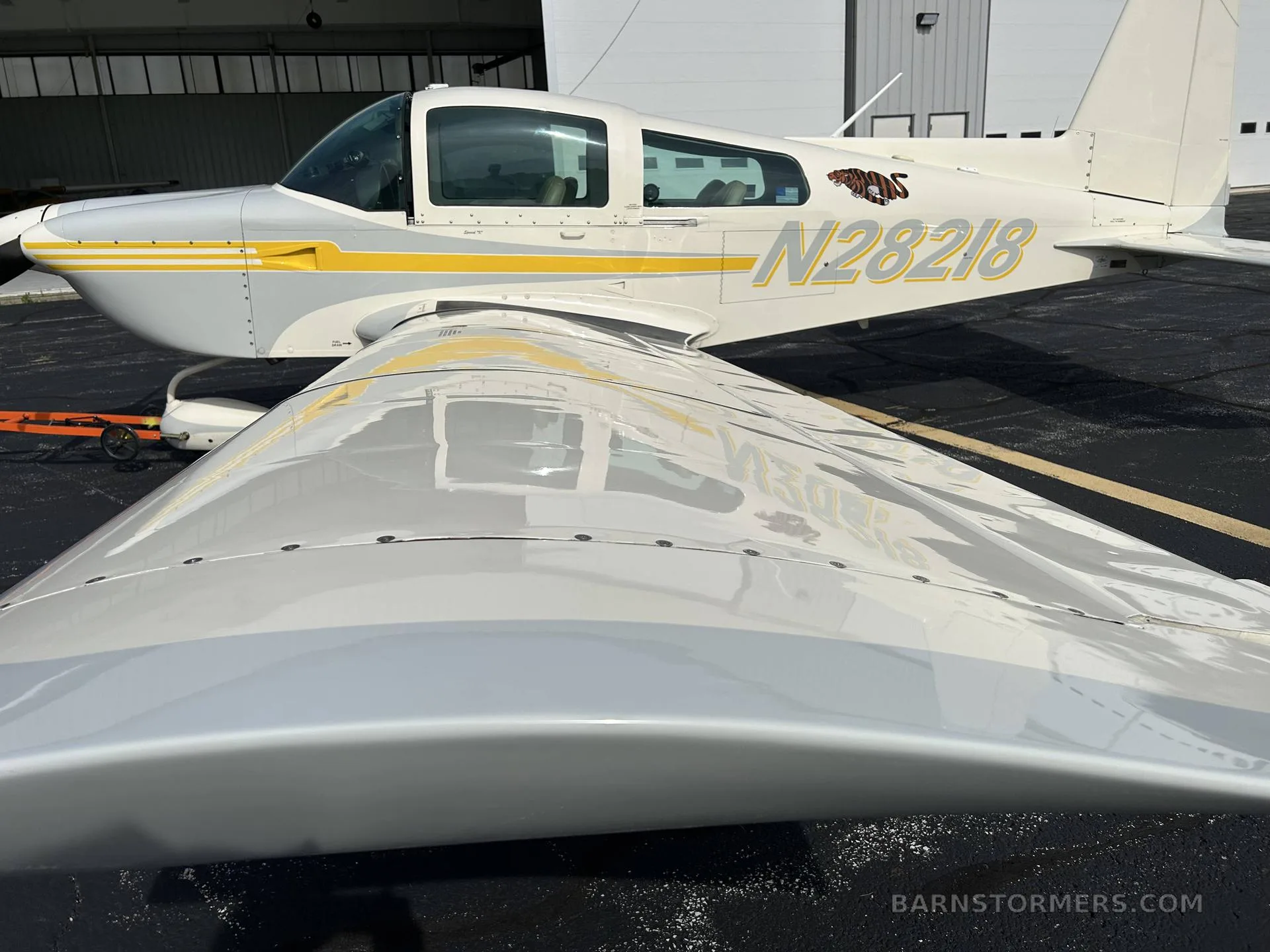 A close-up view of the wing and fuselage of a white and yellow Grumman AA-5B Tiger aircraft with tail number N28218, parked in front of a hangar.