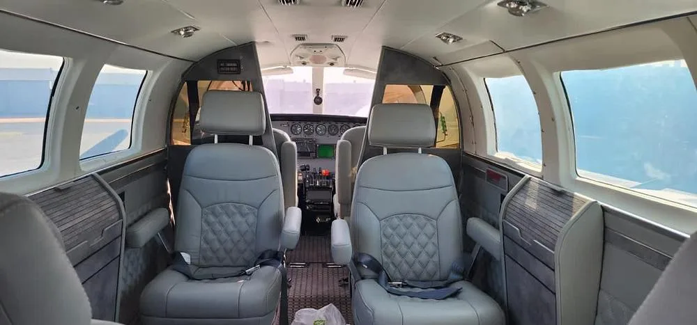 The aircraft cabin interior features light gray quilted leather seats and wood-paneled side walls, looking towards the cockpit.