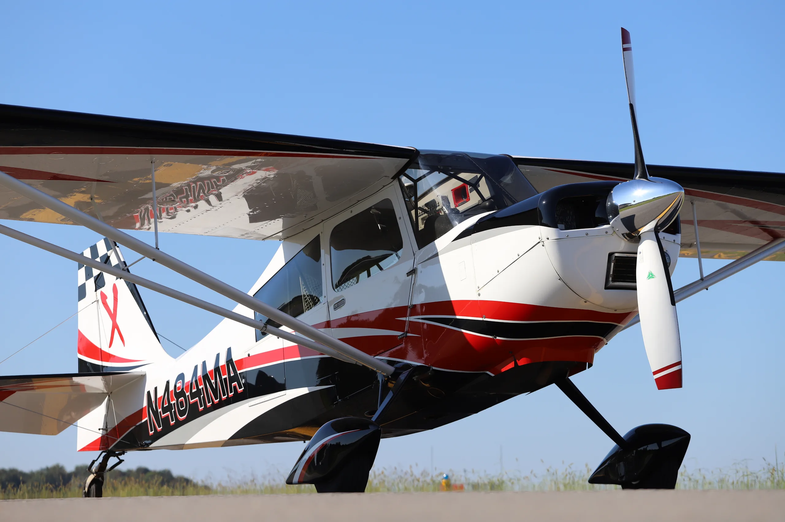 A white, red, and black single-engine taildragger aircraft (N484MA) with a checkered tail, parked on a tarmac under a clear blue sky.