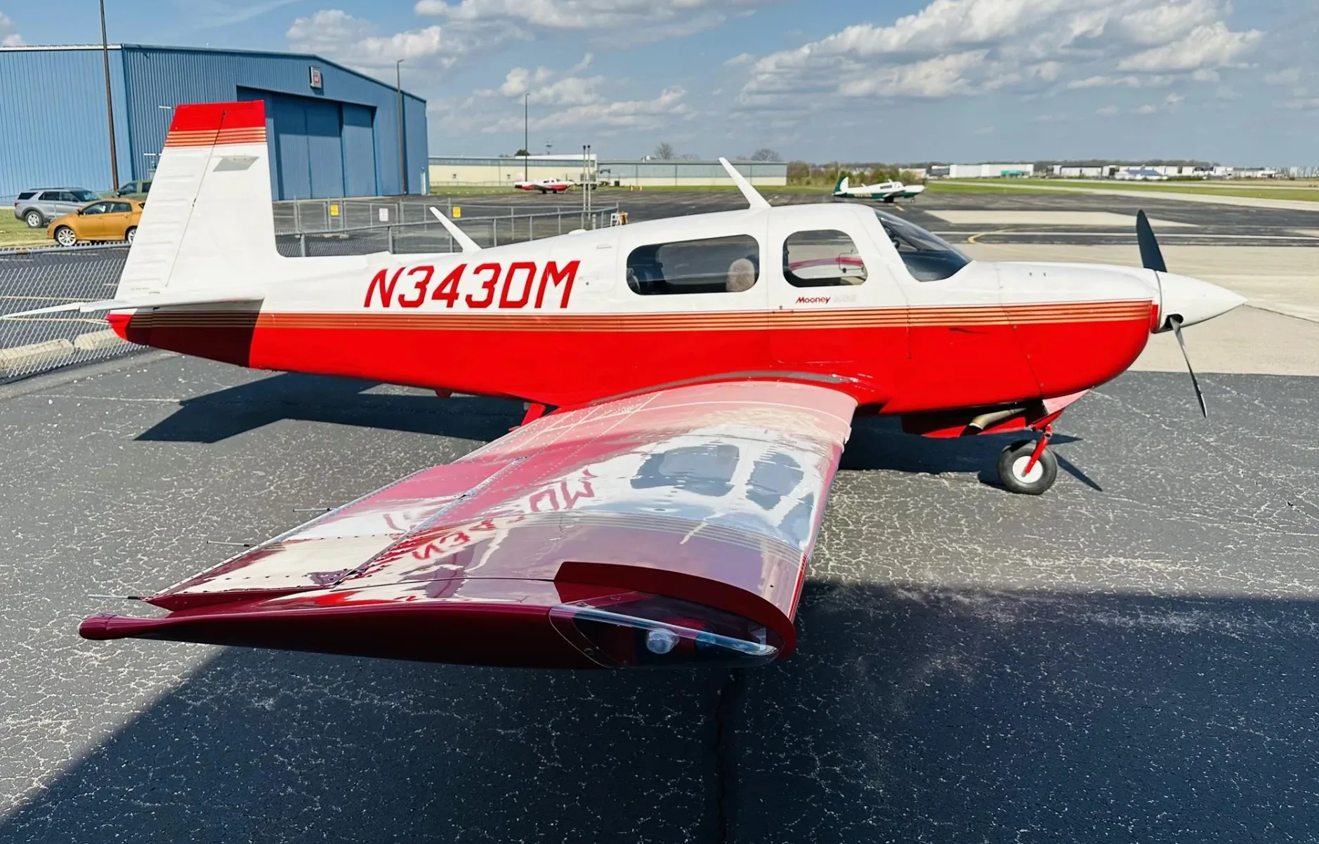 A red and white Mooney aircraft (N343DM) parked on a tarmac with hangars in the background.