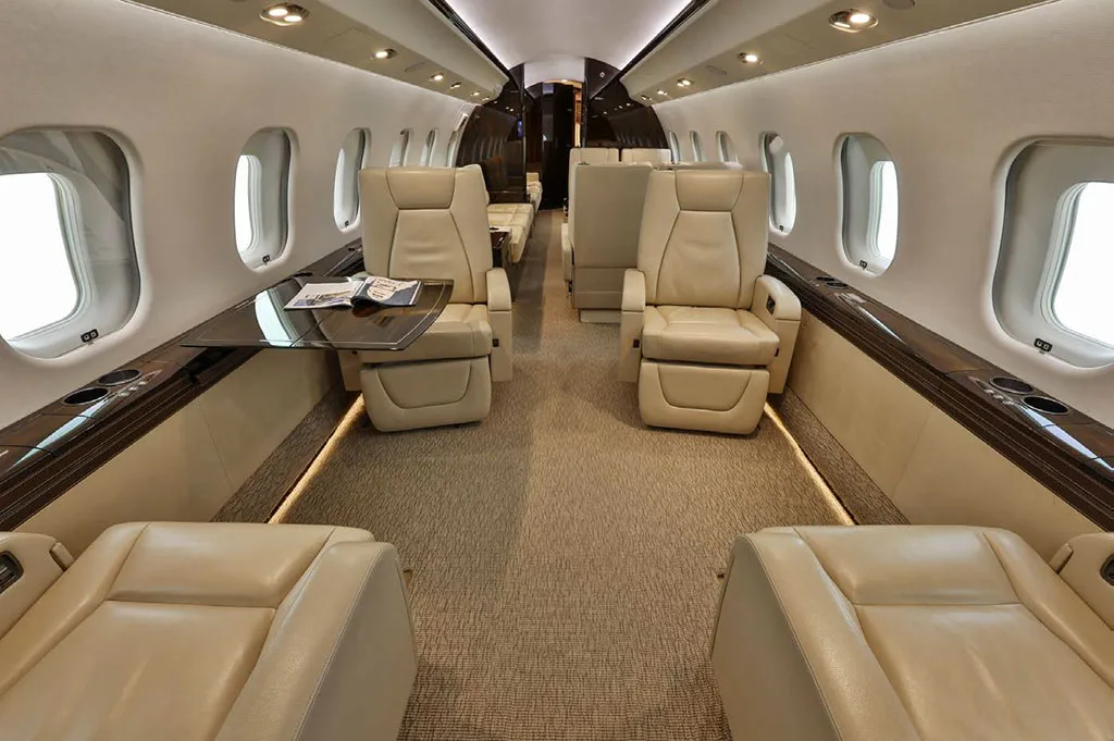 The luxurious cabin interior of a business jet, featuring beige leather seats, a table, and large windows.