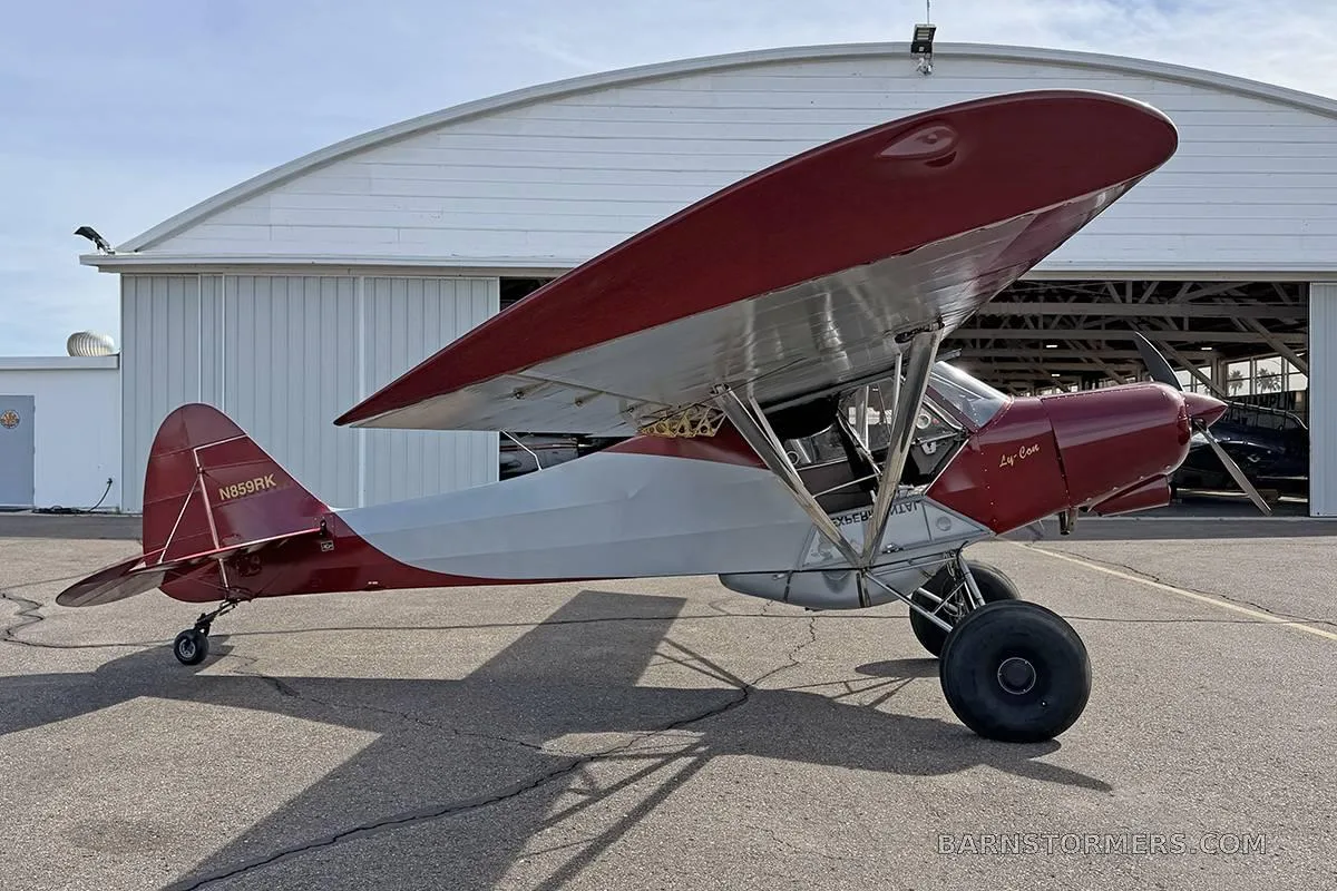 A red and grey high-wing aircraft with large tires, seen from the left side in front of a hangar.