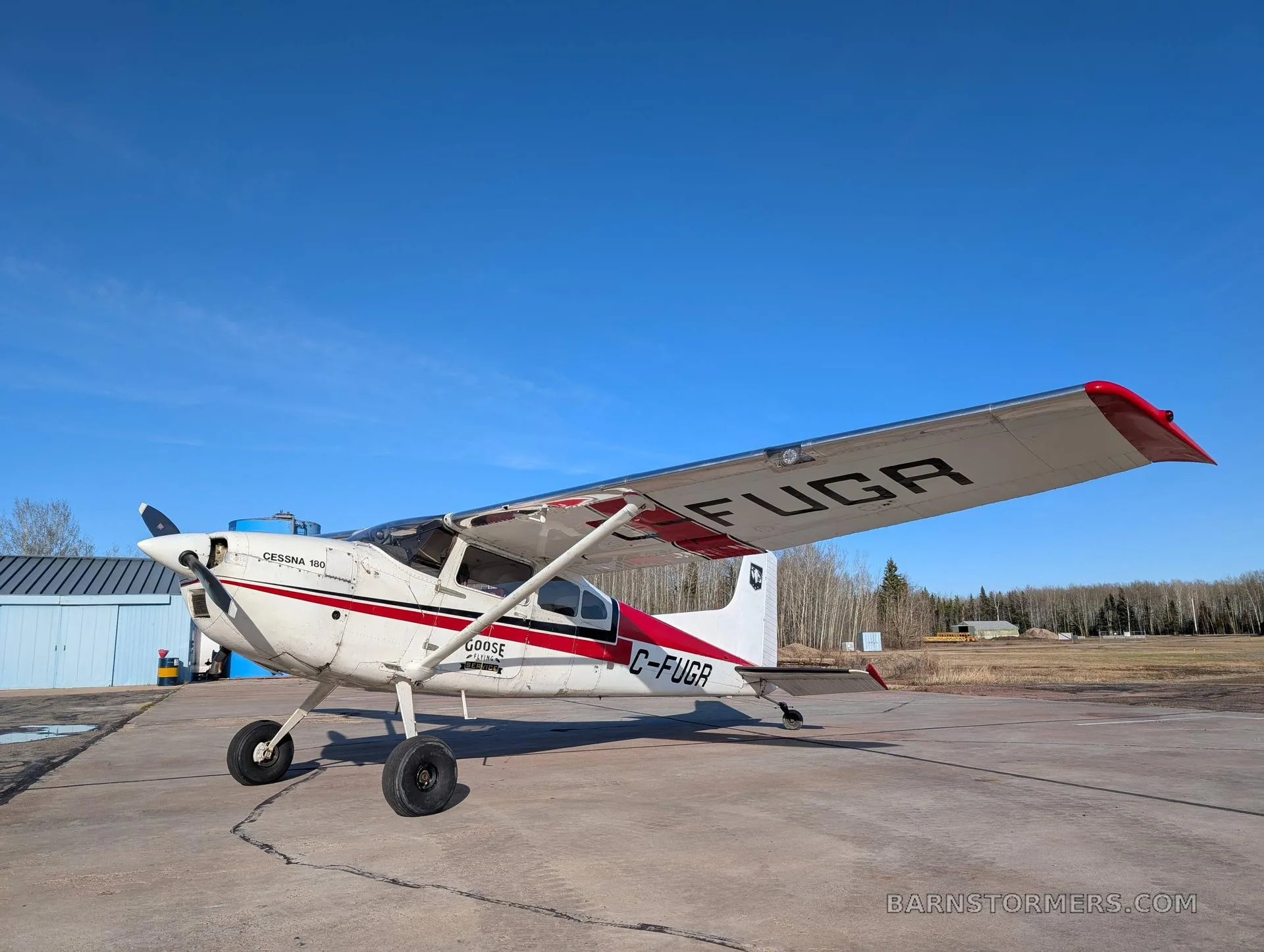 A white and red Cessna 180 (C-FUGR) is parked on a concrete tarmac next to a blue hangar under a clear blue sky.
