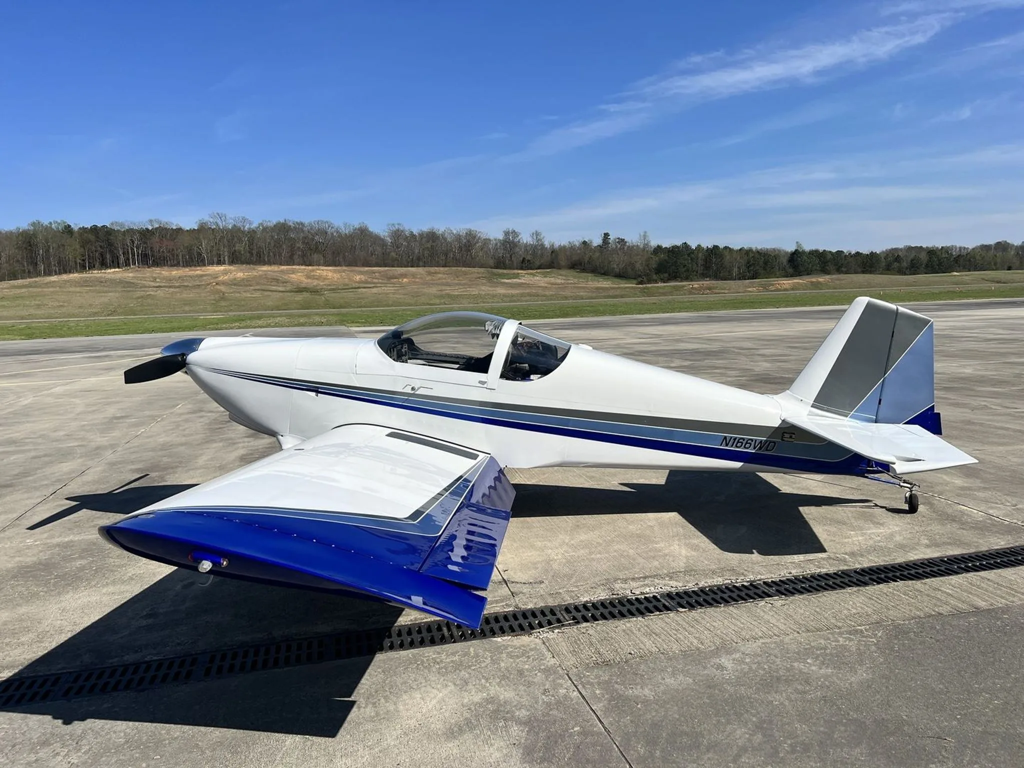 A white Van's RV-6 aircraft with blue and gray stripes parked on an airfield under a clear blue sky.