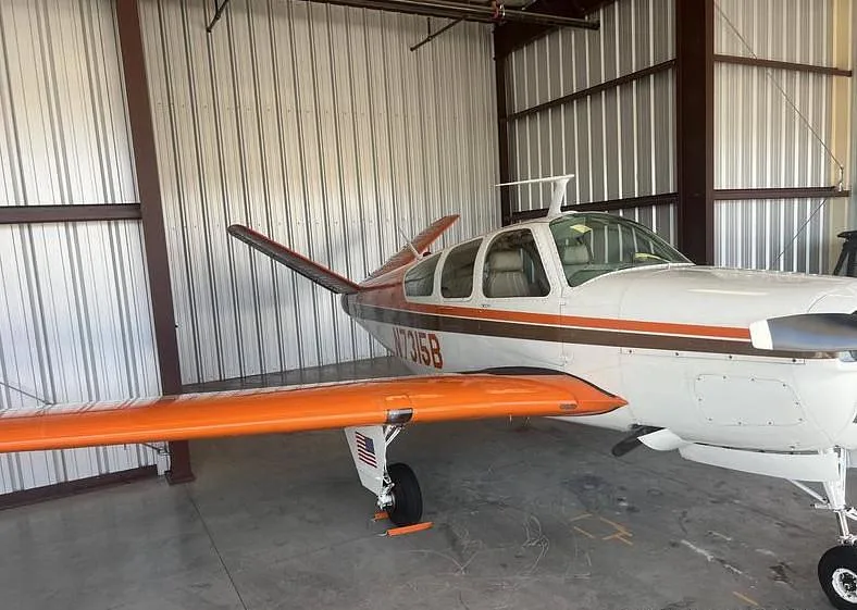 A white and brown Beechcraft Bonanza N7315B with orange wings parked in a hangar, viewed from the side.
