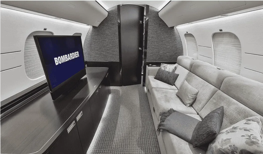 A private jet cabin interior with a long sofa, a TV displaying "BOMBARDIER", and dark textured walls.