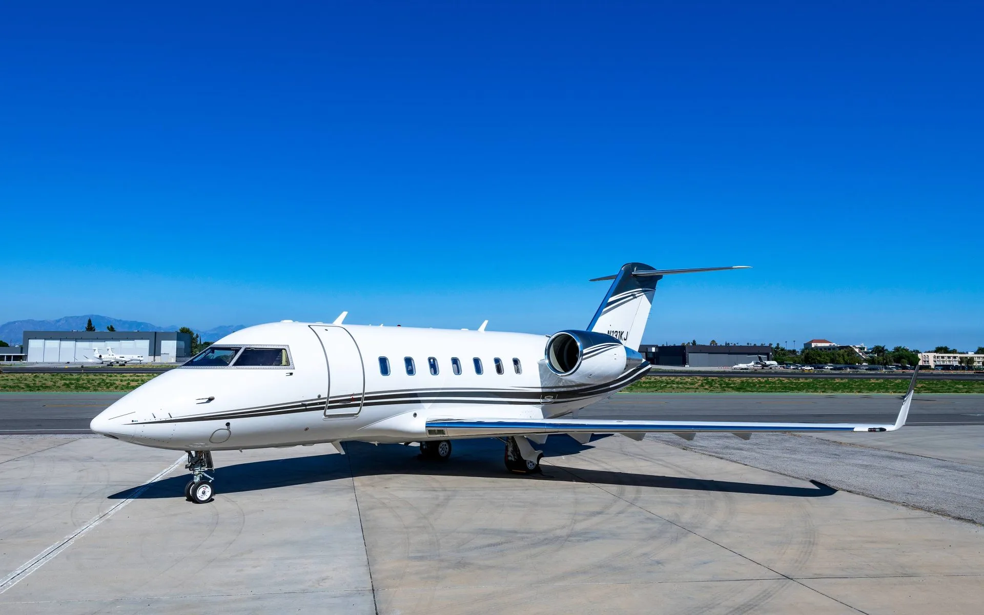 A white Bombardier Challenger business jet with dark and white stripes is parked on an airport tarmac under a clear blue sky.