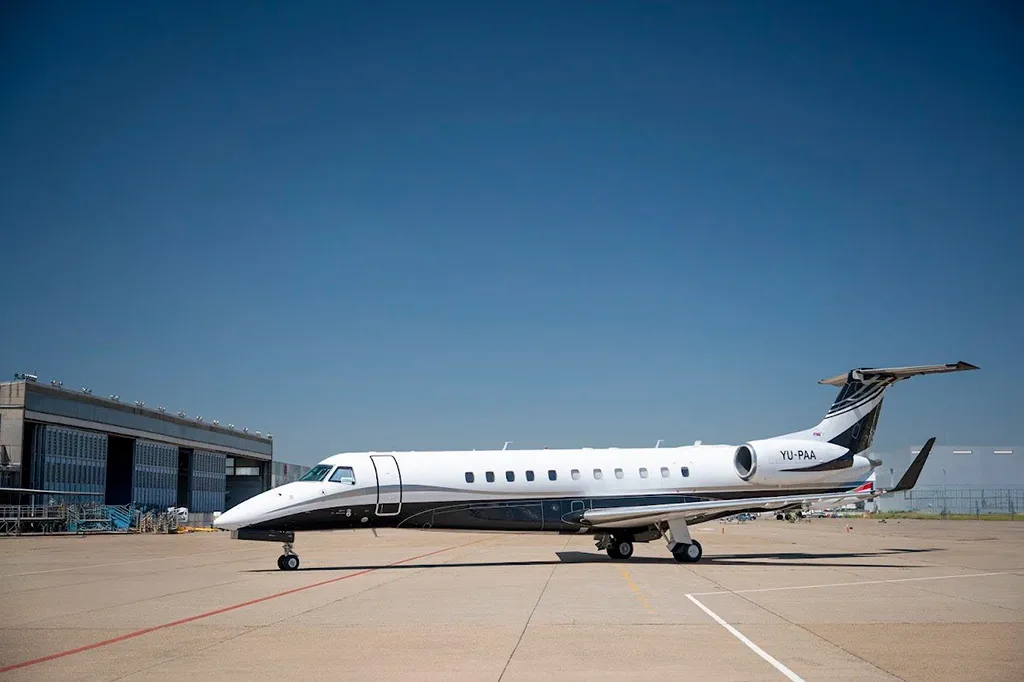 A white and black Embraer Legacy 600 business jet with registration YU-PAA parked on a tarmac next to a hangar.