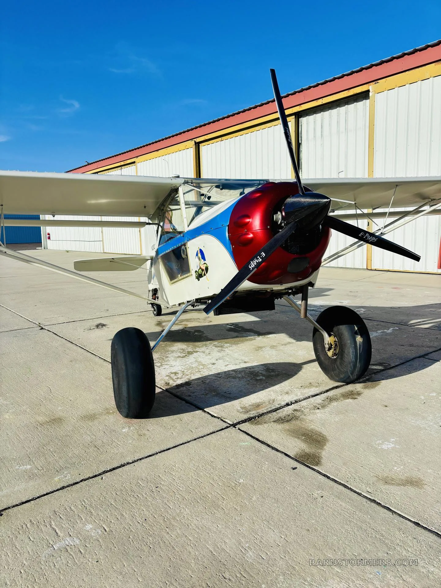 A white Kitfox aircraft with a red and blue cowl, large tires, parked on a tarmac in front of hangars.