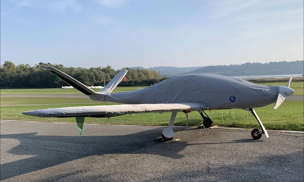 A light gray covered aircraft with its propeller exposed is parked on an airfield next to a body of water.