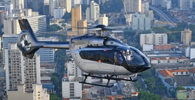 A silver and black Airbus H145 helicopter with two pilots flies over a dense city landscape with tall buildings.