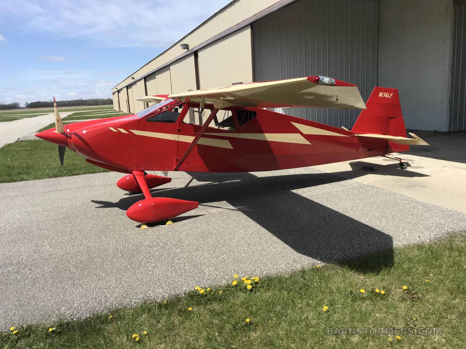 A red and cream single-engine aircraft with floats parked on an airfield next to a hangar.