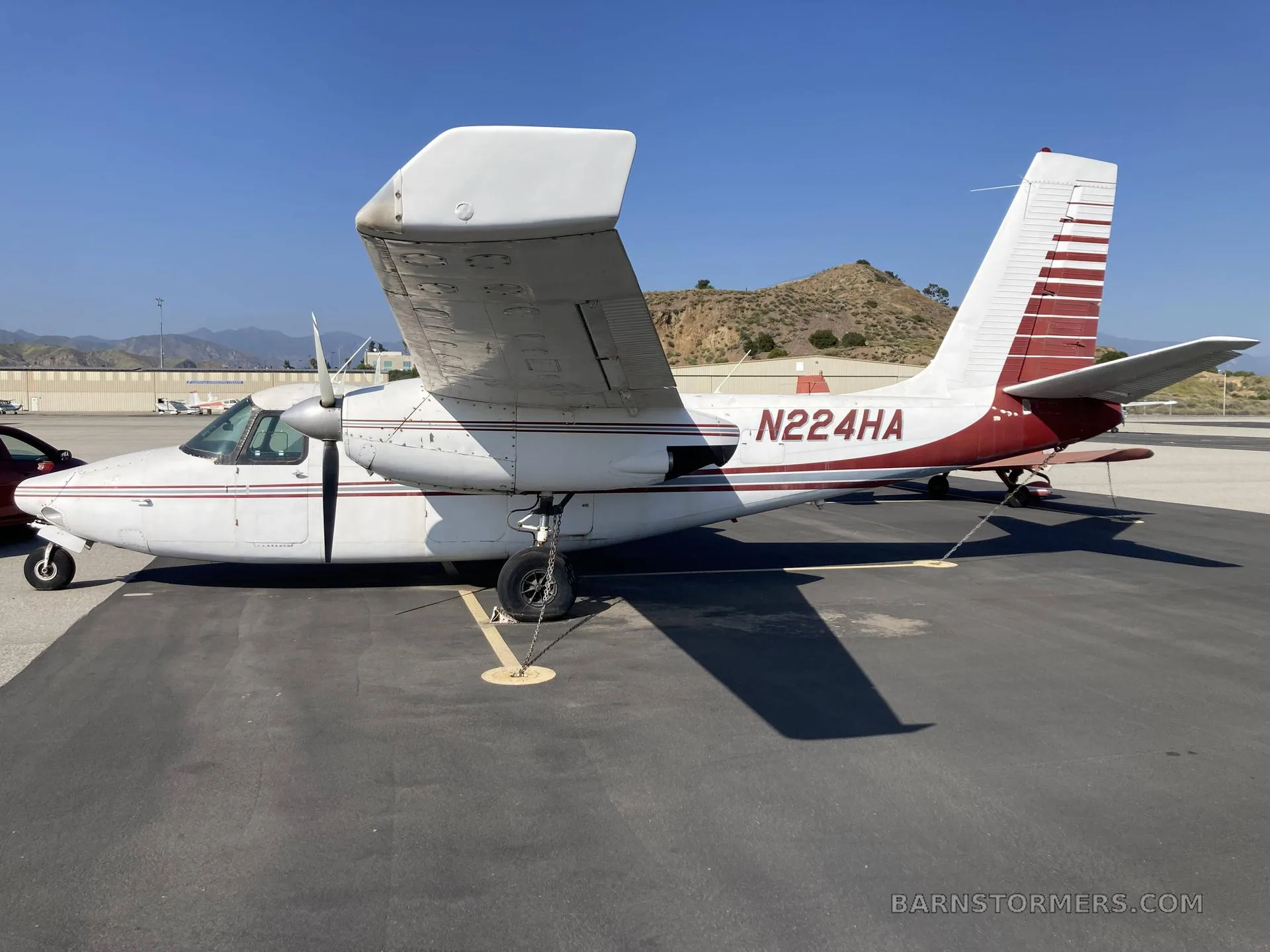 A white and red twin-engine Commander aircraft (N224HA) parked on an asphalt tarmac with mountains in the background.