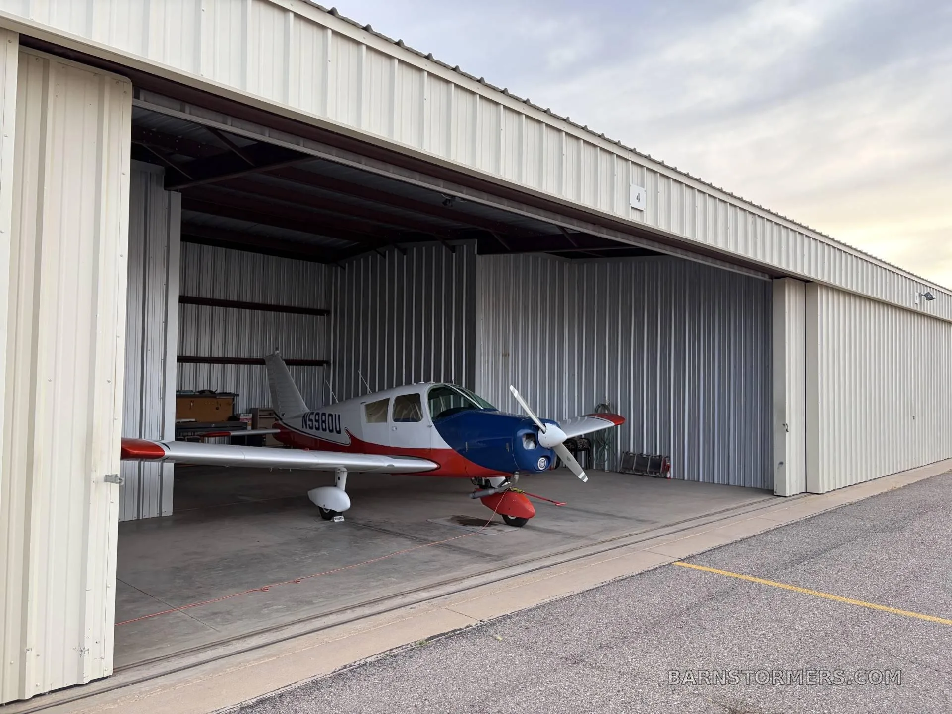 A red, white, and blue single-engine propeller aircraft (N5980U) is partially pulled out of a hangar.