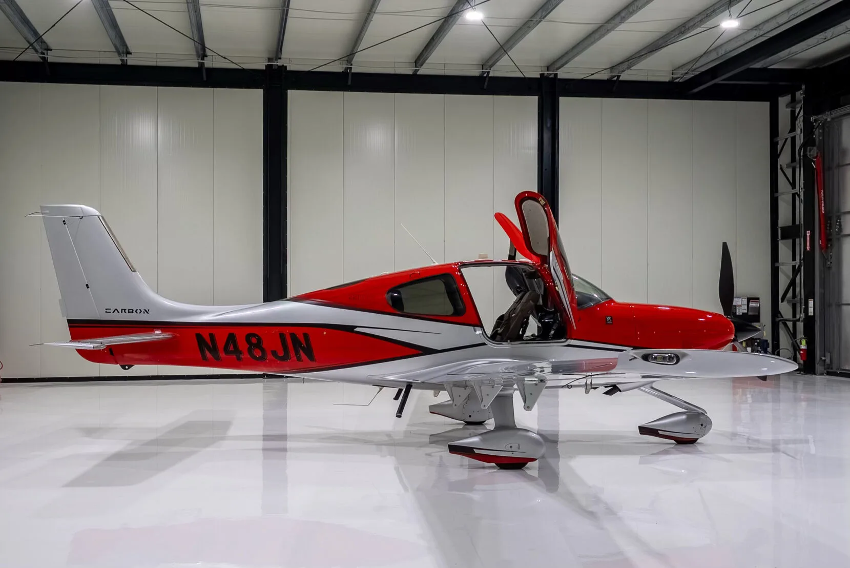 Side view of a red and silver Cirrus SR22T Carbon aircraft with registration N48JN and open doors, in a hangar.