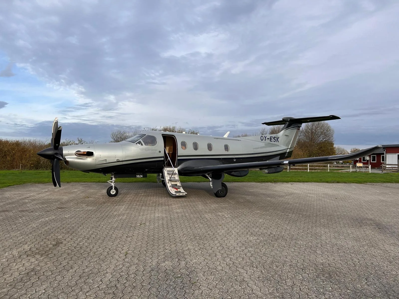 A silver and black Pilatus PC-12 turboprop aircraft with its door open and stairs extended, parked on a paved surface under a cloudy sky.