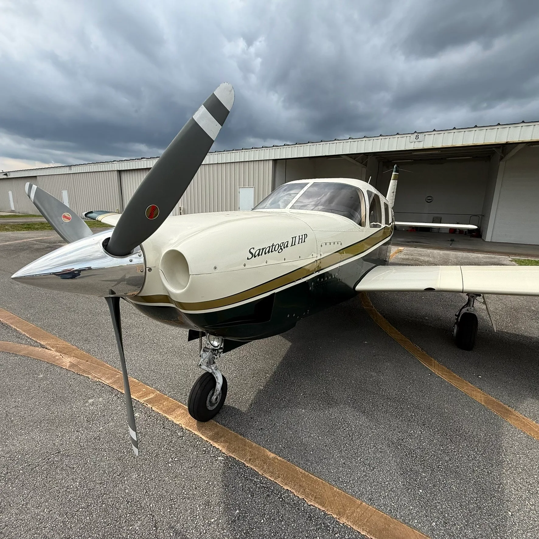 A Piper Saratoga II HP, N221AH, with a white, gold, and dark green paint scheme, parked on an airport tarmac under a cloudy sky.