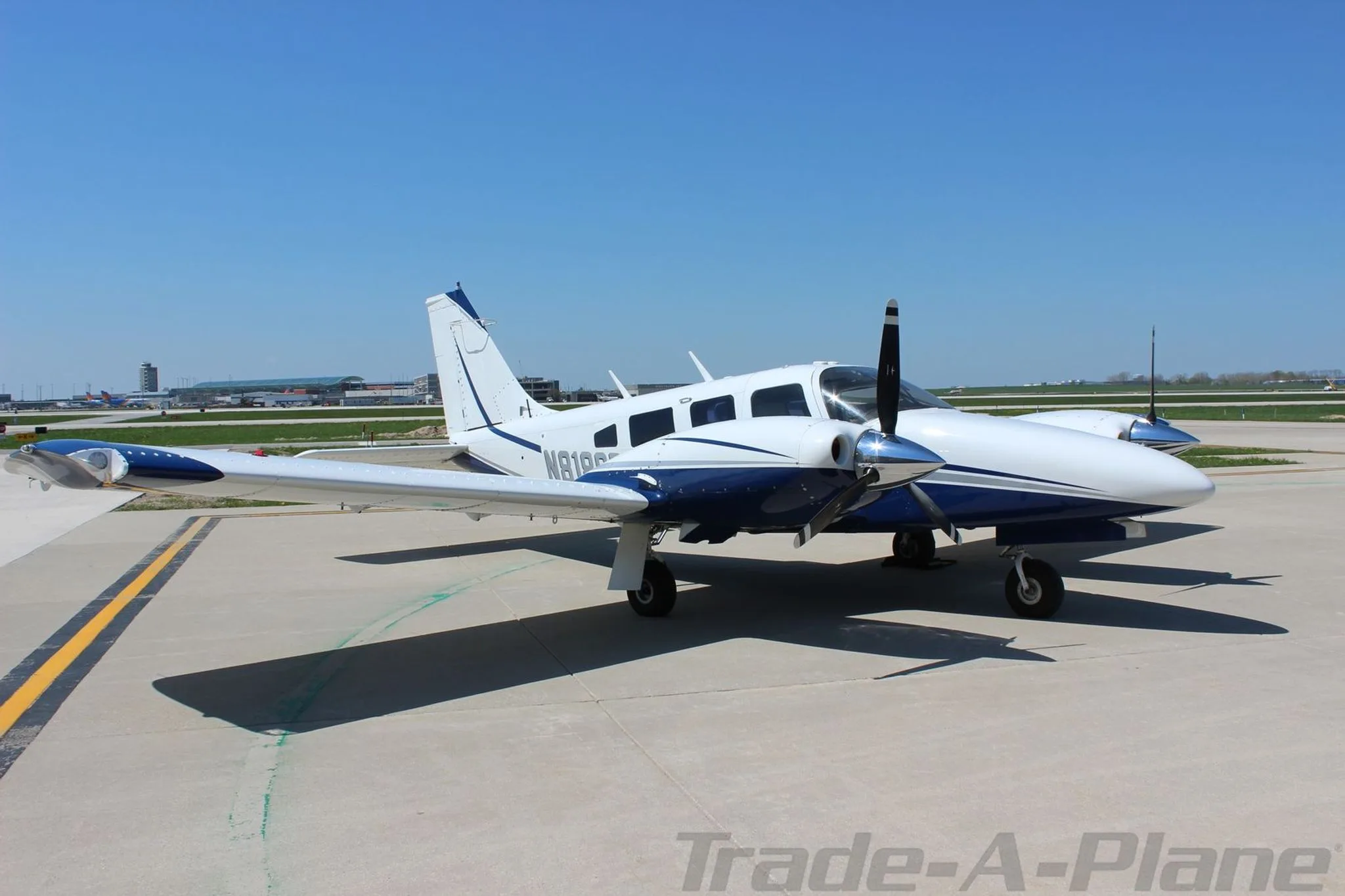 A side view of the white and blue Piper Seneca aircraft N8186R with its propellers visible, parked on an airport tarmac.