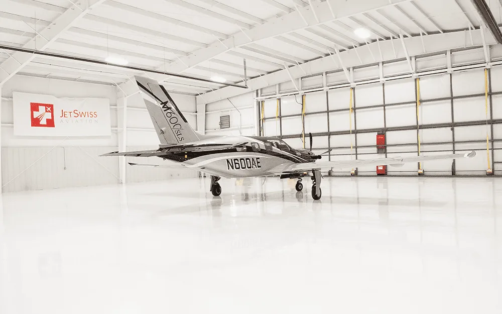 A Piper M600/SLS aircraft (N600AE) with a black and silver paint scheme is parked in a clean, white hangar, viewed from the front-left.