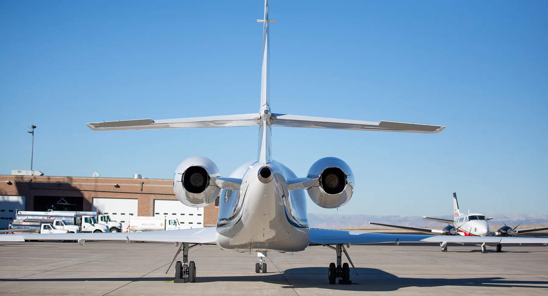 A rear view of a white twin-engine business jet parked on an airfield with other aircraft in the background.