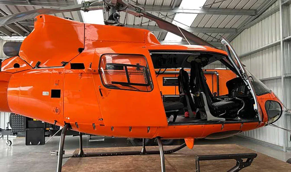 A side view of a bright orange helicopter with its doors open, revealing the black seats inside, parked in a hangar.