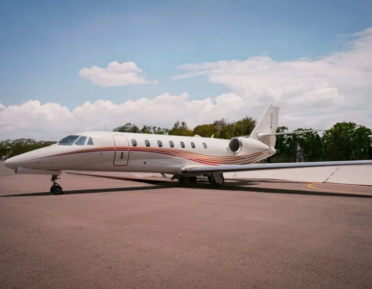 A white private jet with red and gold stripes on its fuselage is parked on an asphalt apron under a partly cloudy sky.