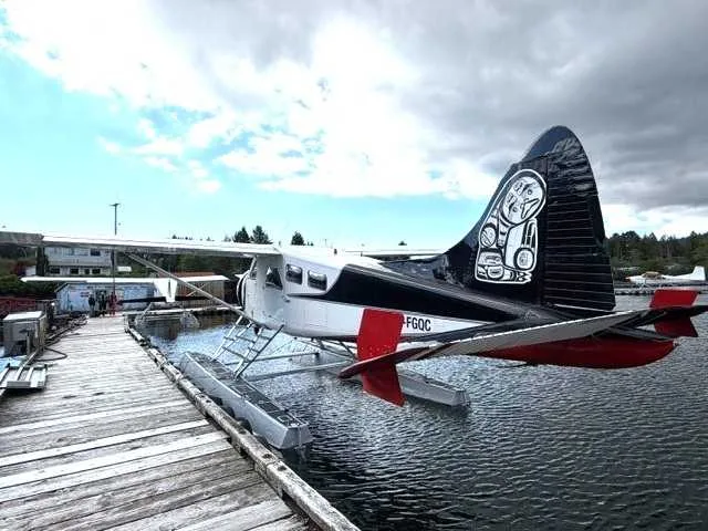 A white and black floatplane with red accents and distinctive tail art, C-FGQC, docked at a wooden pier on a cloudy day.