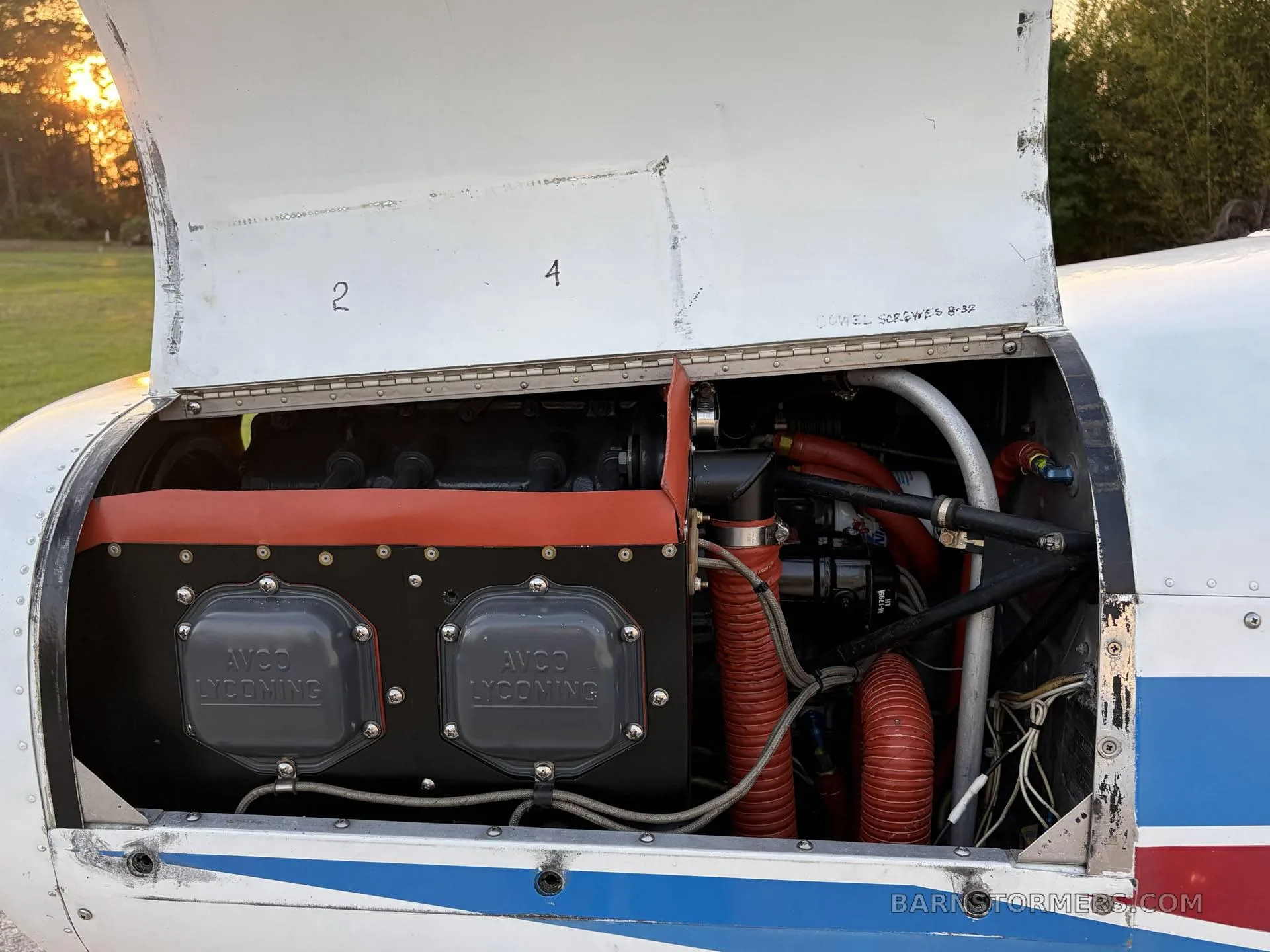 The open engine cowling of a white aircraft reveals an Avco Lycoming engine with red hoses and components.