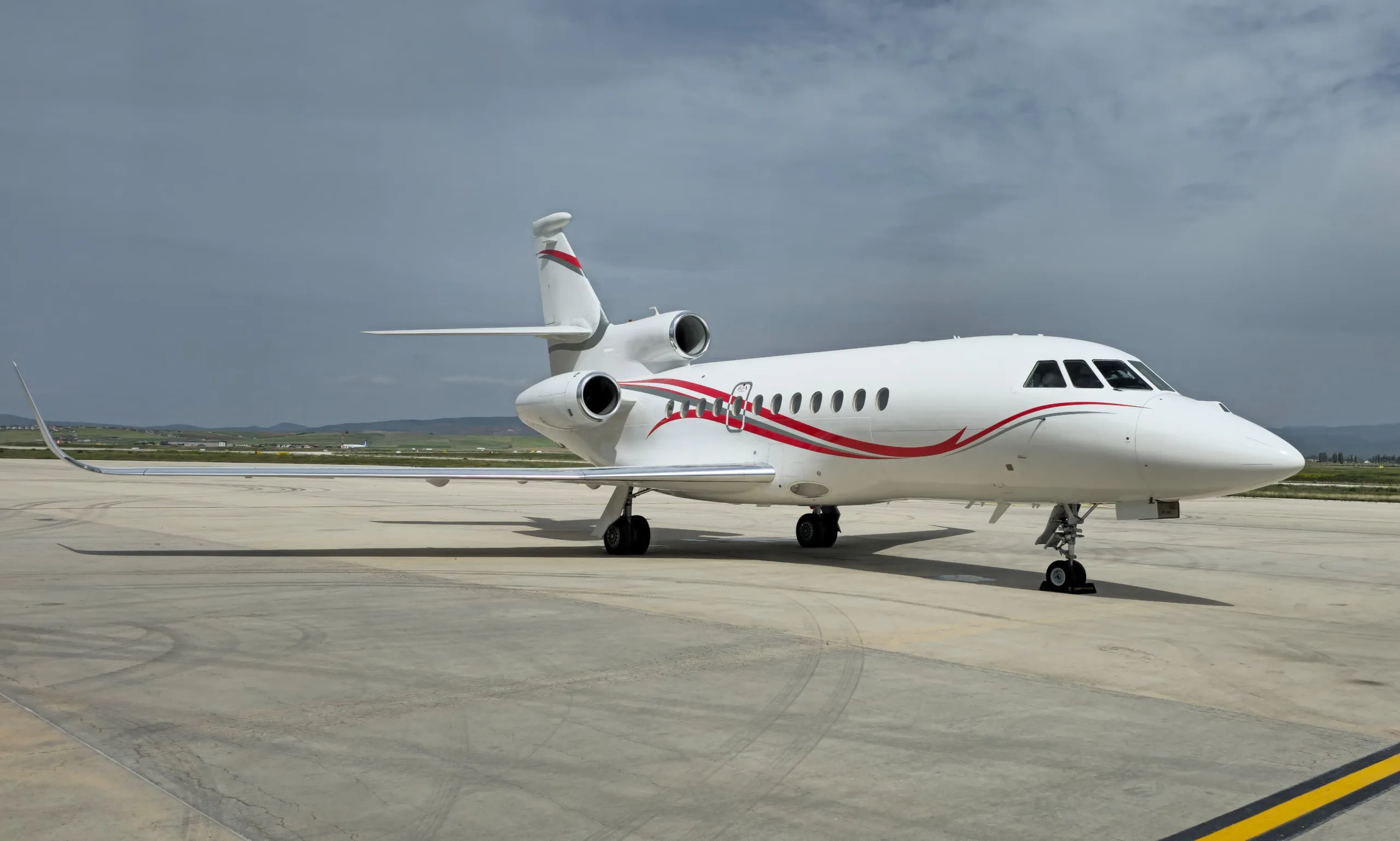A white business jet with red and grey stripes is parked on an airfield under a cloudy sky.