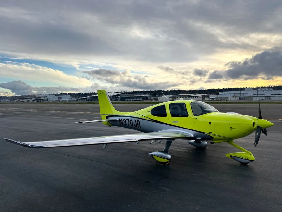 A bright yellow and silver Cirrus SR22 aircraft with registration N370JR parked on an airport tarmac with a cloudy sunset sky.