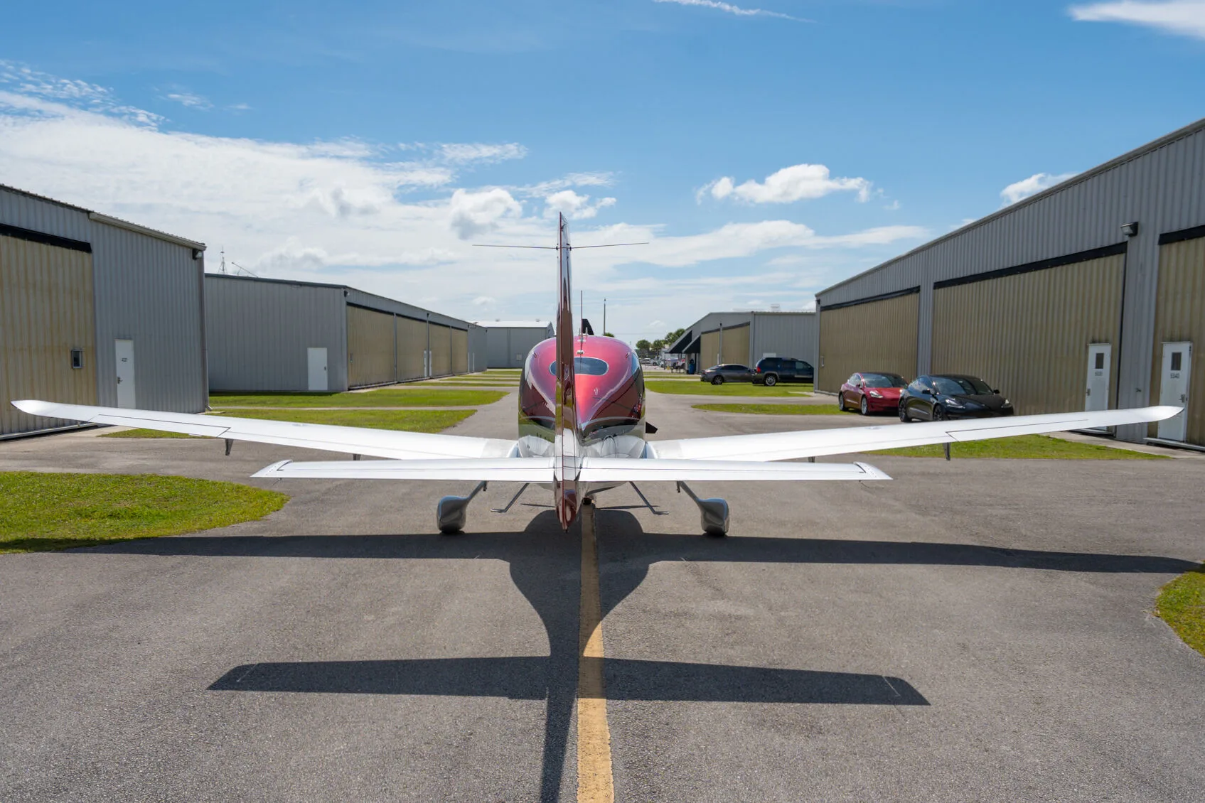Rear view of a red and silver Cirrus SR22 GTS parked on an airfield between hangars.