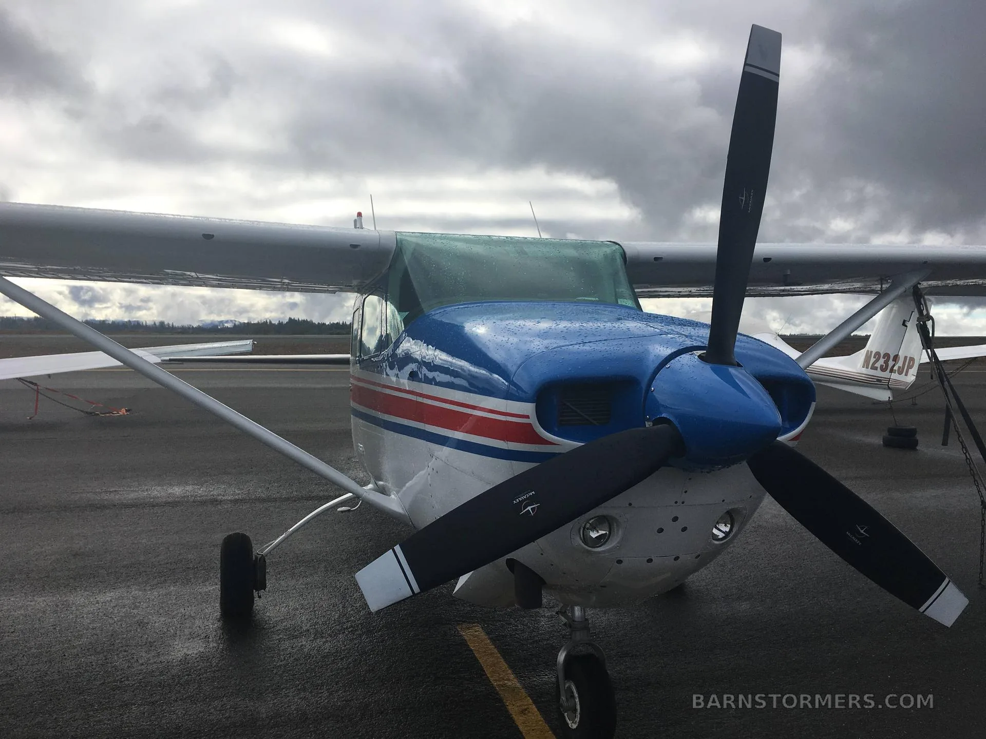 A front view of a white, red, and blue Cessna Stationair 6 II with a propeller, parked on a wet tarmac under a cloudy sky.