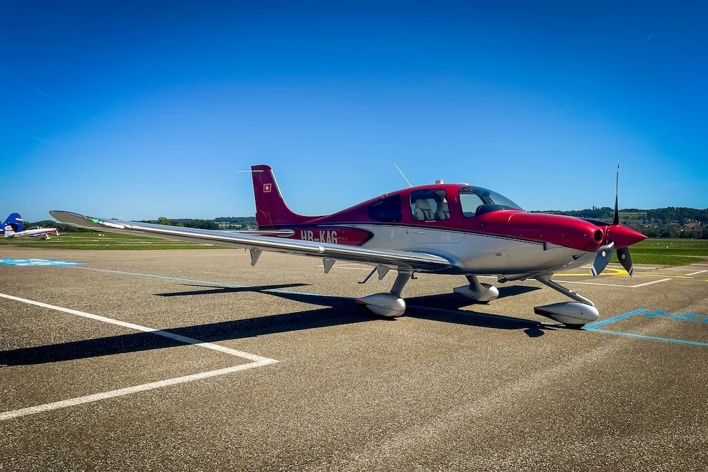 Three-quarter front view of a red and silver Cirrus SR22 aircraft parked on an airfield under a clear blue sky.