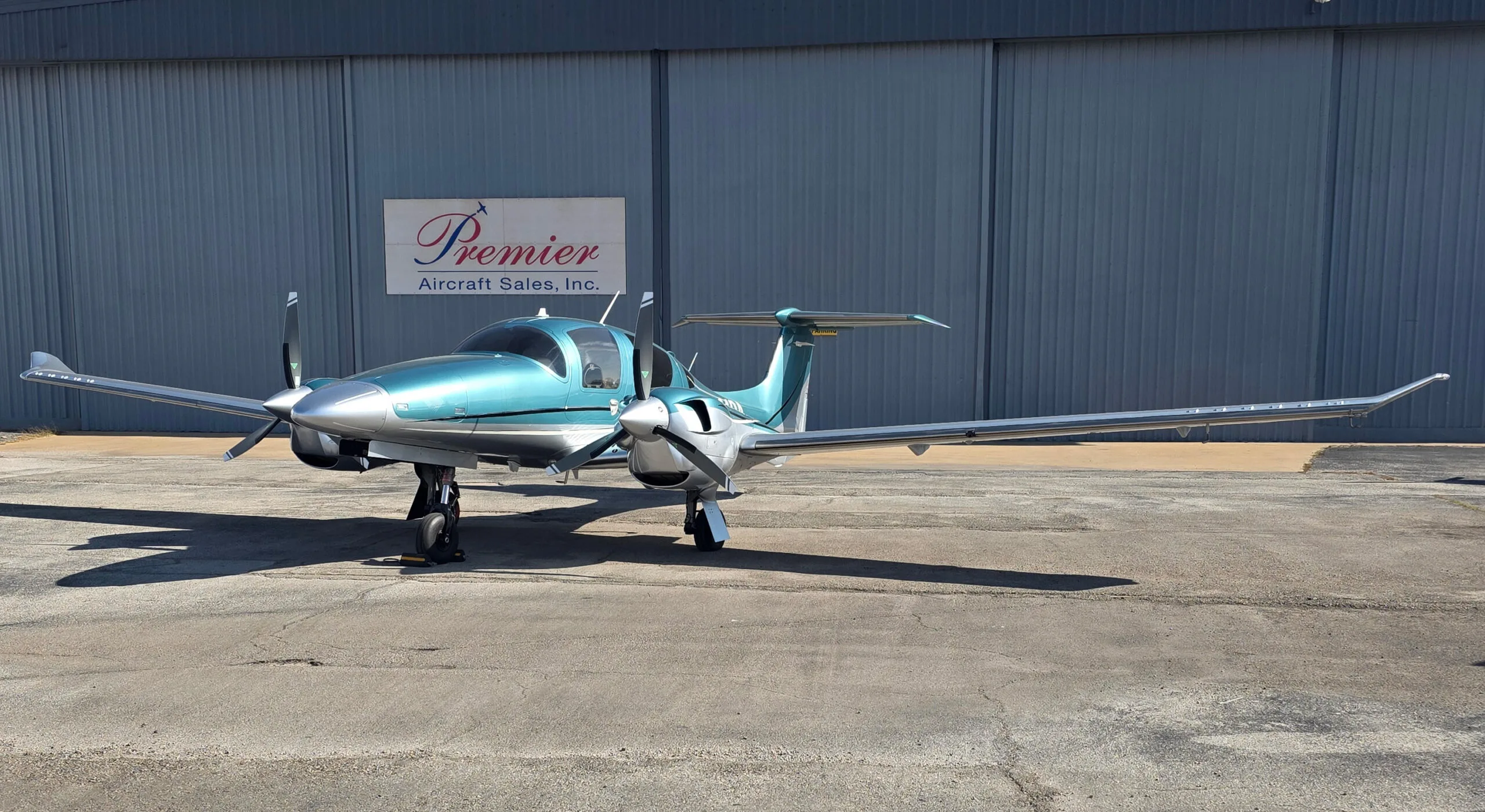 A teal and silver Diamond DA62 twin-engine aircraft parked on an asphalt ramp in front of a hangar.