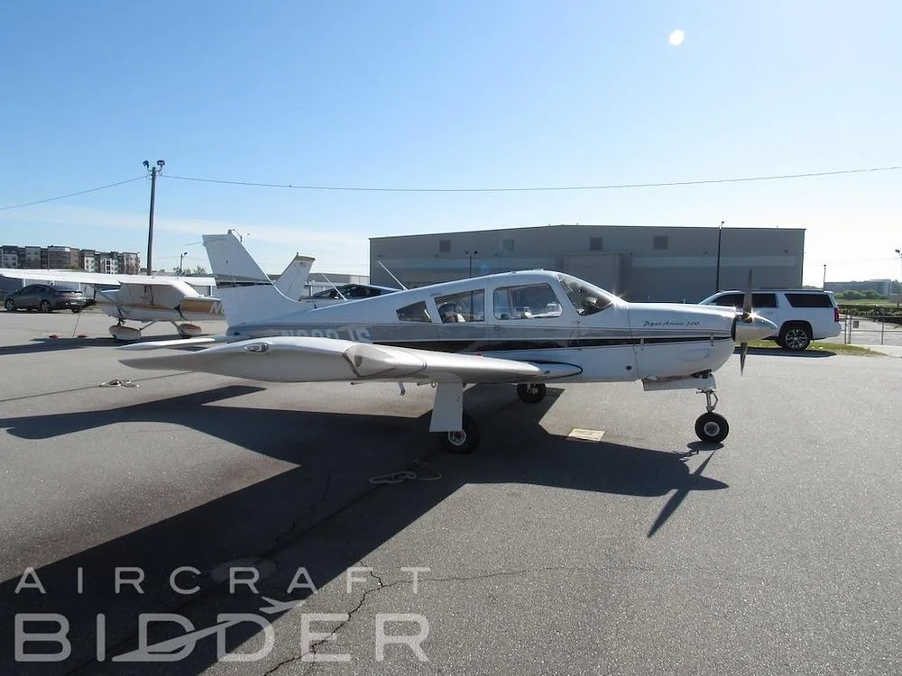 A side view of the white Piper Arrow 200 (N822JS) with black and grey stripes, parked on an asphalt tarmac.
