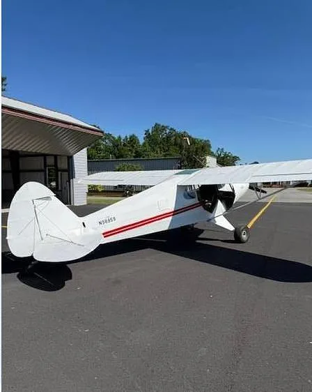 A three-quarter rear view of a white aircraft with red wings and stripes, showing the N-number N98959, parked on an asphalt tarmac.