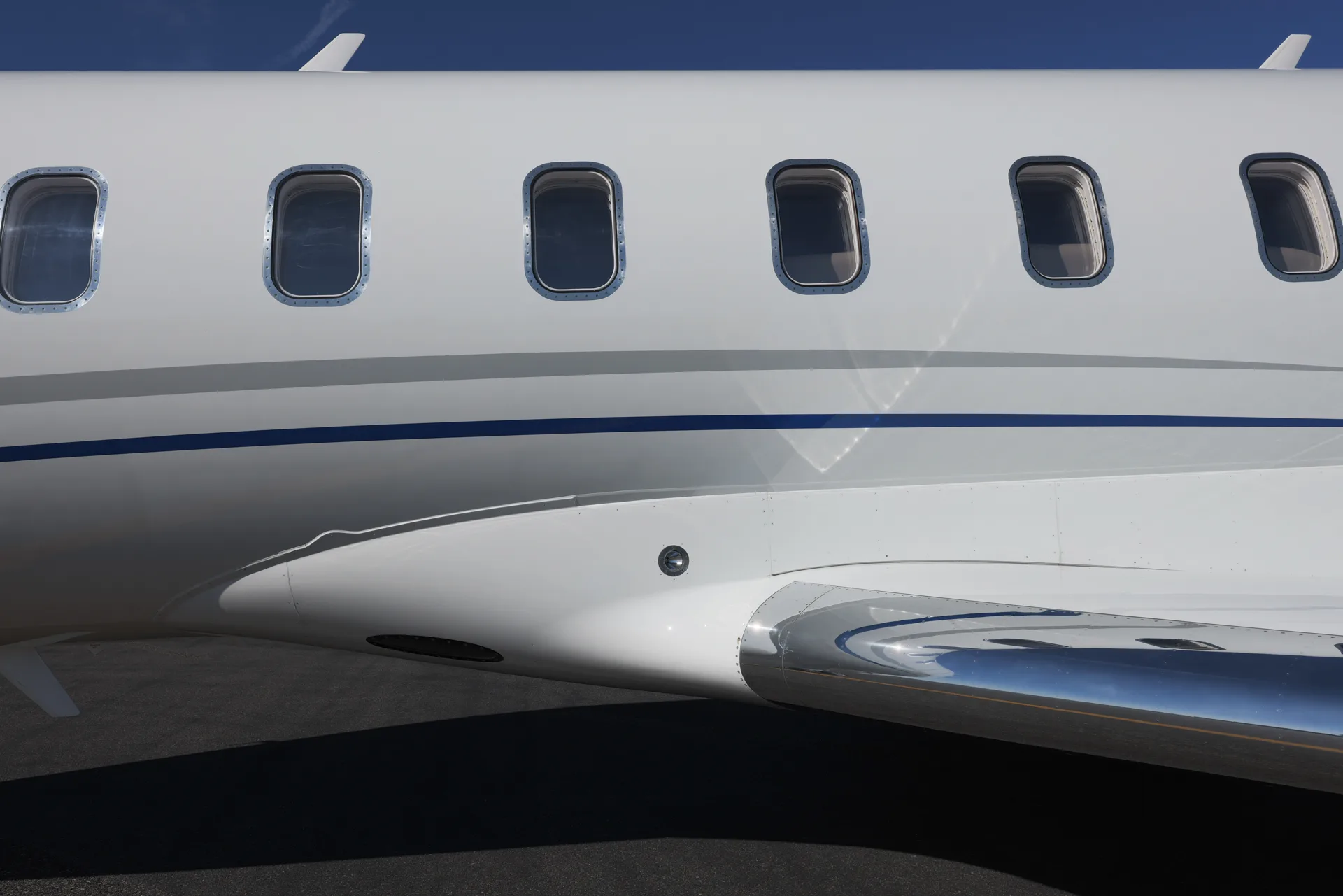 A close-up of the white fuselage and polished wing of a business jet, showing cabin windows and a blue stripe.