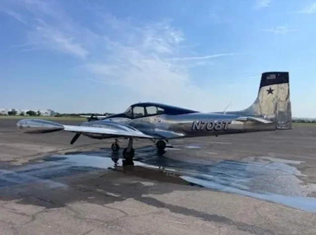A silver and blue twin-engine aircraft, N708T, parked on a wet tarmac under a clear sky.