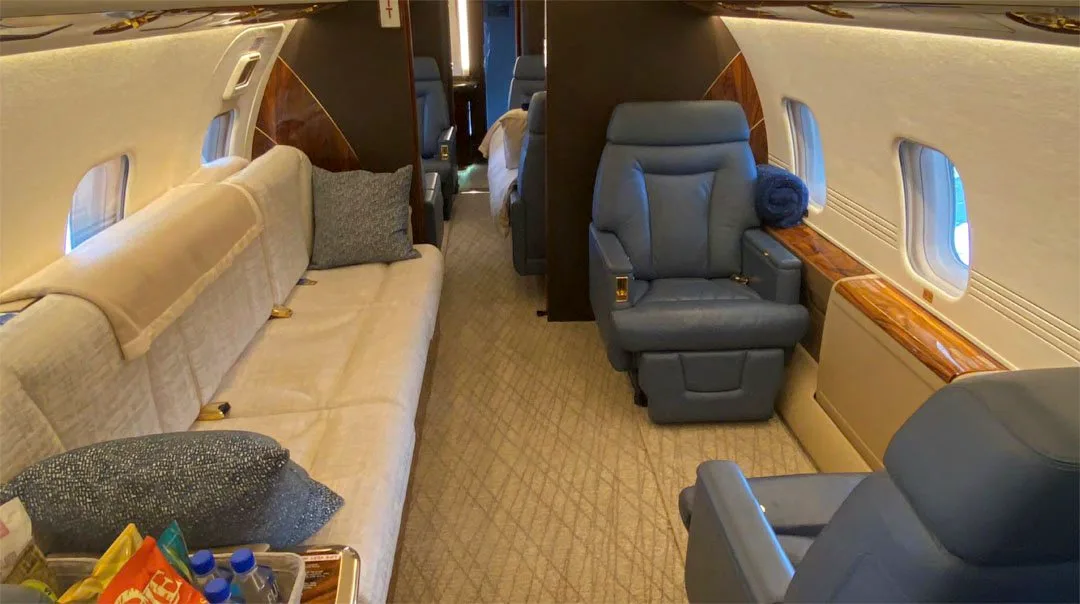 The luxurious cabin interior of a business jet features a beige sofa, blue leather seats, and wood accents.