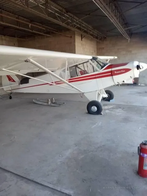 A white Piper Super Cub with red stripes is parked inside a hangar, with aircraft skis on the ground nearby.