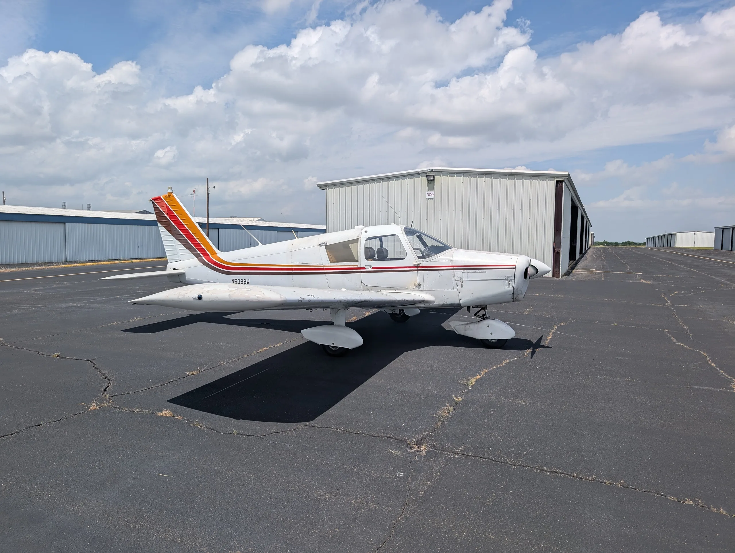 A white Piper Cherokee aircraft with brown and orange stripes, parked on an asphalt apron with hangars in the background.