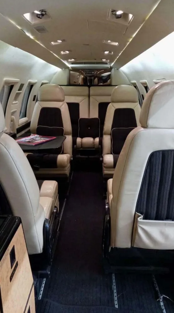 View of a private jet cabin with beige and dark brown leather seats, a dark carpet, and overhead lighting.