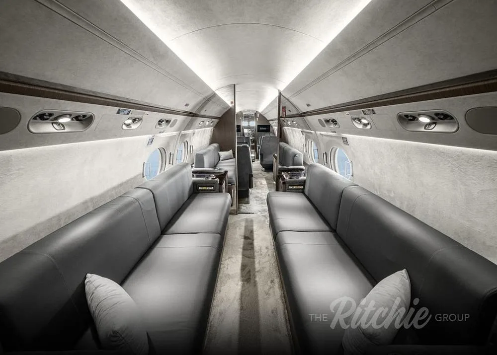 The spacious interior cabin of a private jet features two long grey leather couches facing each other along the aisle.