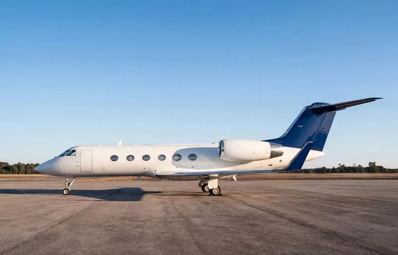 A white business jet with a dark blue tail is parked on an asphalt runway under a clear blue sky.