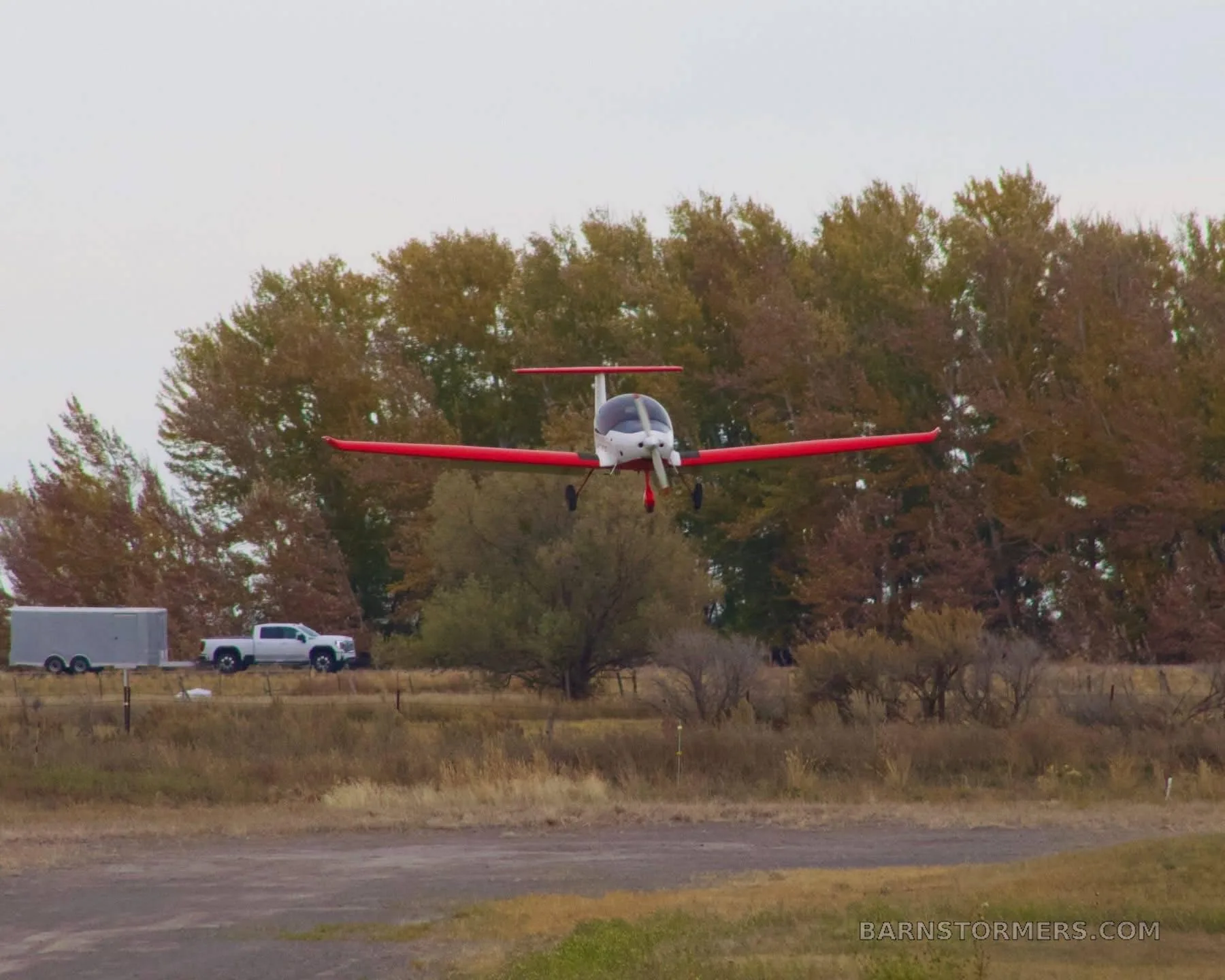 A small white and red experimental aircraft, possibly an ATEC Zephyr, is seen in flight against a backdrop of autumn trees and a rural landscape.