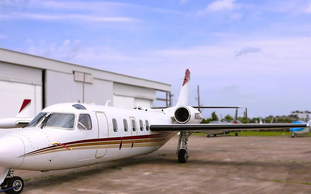 A white business jet with red and gold stripes, registration N959JJ, parked on an airfield next to a hangar.