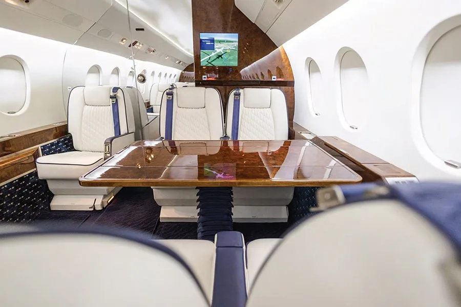 A private jet cabin with white and blue seats facing a large polished wooden table.
