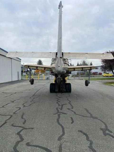 The rear view of a bare metal aircraft fuselage, missing wings and engine, is being moved by a yellow forklift on an asphalt tarmac.
