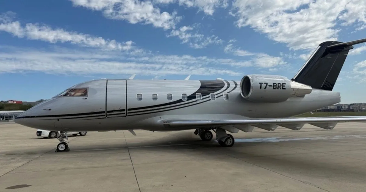 A silver private jet with black stripe livery and registration T7-BRE parked on an airfield under a blue sky.