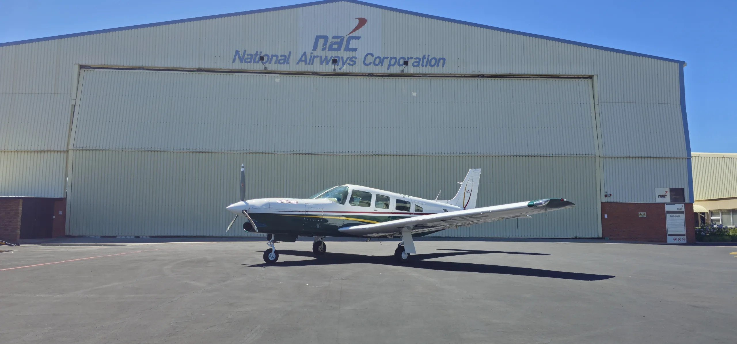 A white and dark green single-engine propeller aircraft with gold stripes is parked on an apron in front of a large hangar under a clear blue sky.